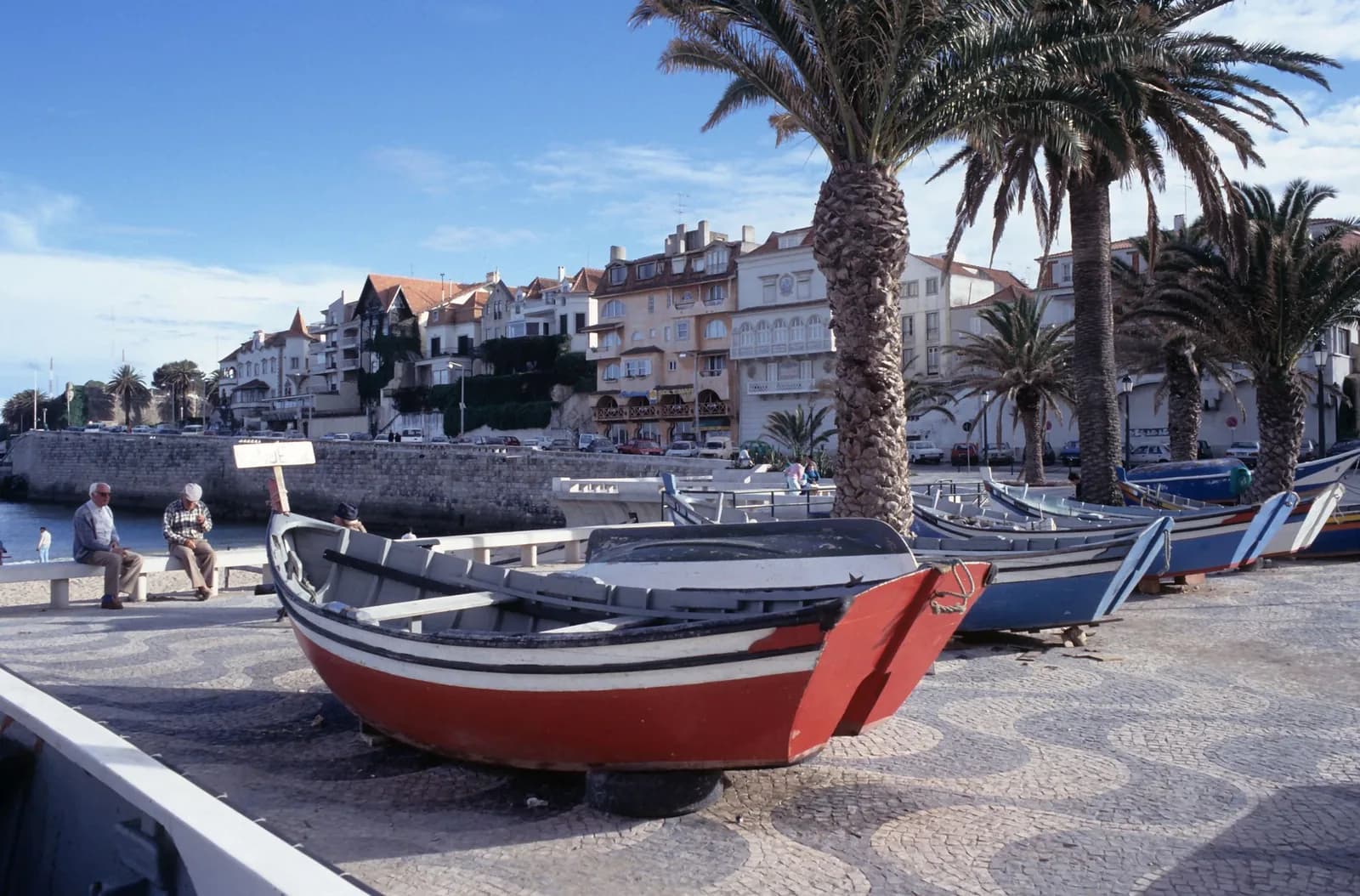 Barques de pêcheurs sur la promenade pavée de Cascais, maisons et palmiers
