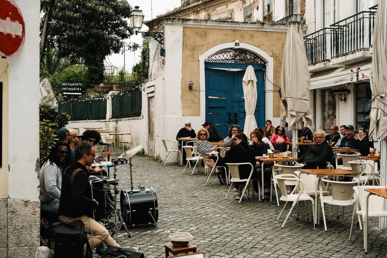Terrasse de café sur une place pavée de Lisbonne, clients attablés sous les parasols et musicien de rue