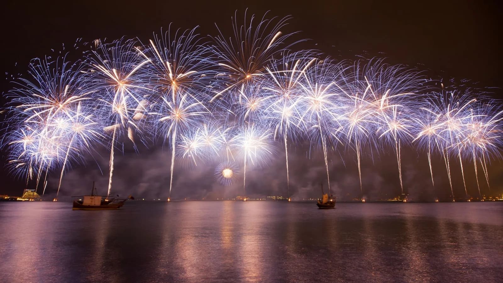 Feu d'artifice nocturne au-dessus d'un fleuve, avec deux voiliers traditionnels et les reflets des lumières sur l'eau