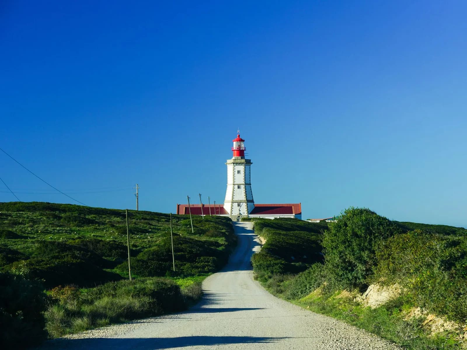 Chemin de terre menant au phare blanc à lanterne rouge du Cabo Espichel, entouré de végétation