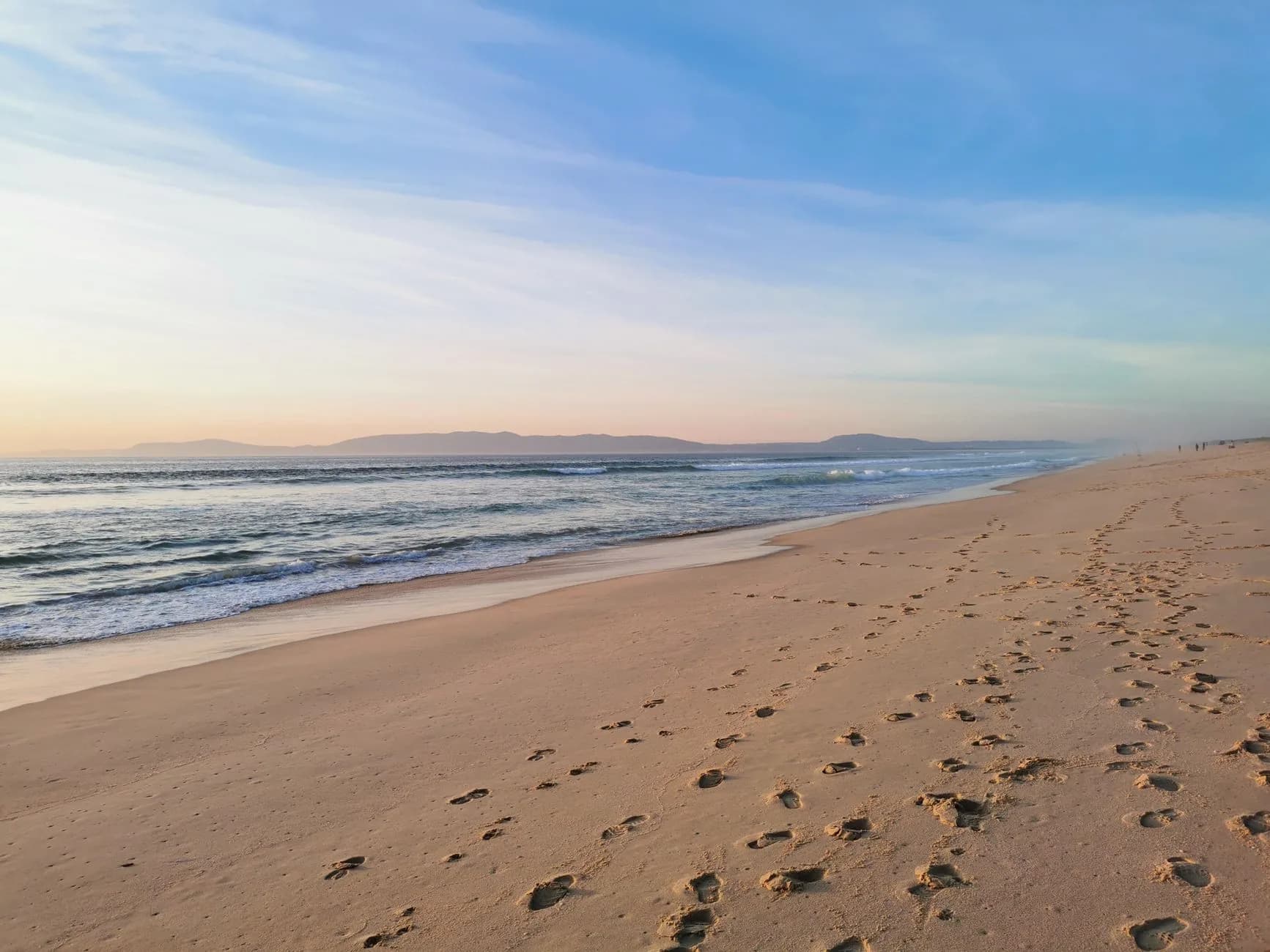Plage de sable doré à marée basse, traces de pas et océan Atlantique sous un ciel pastel, sur la côte de l'Alentejo