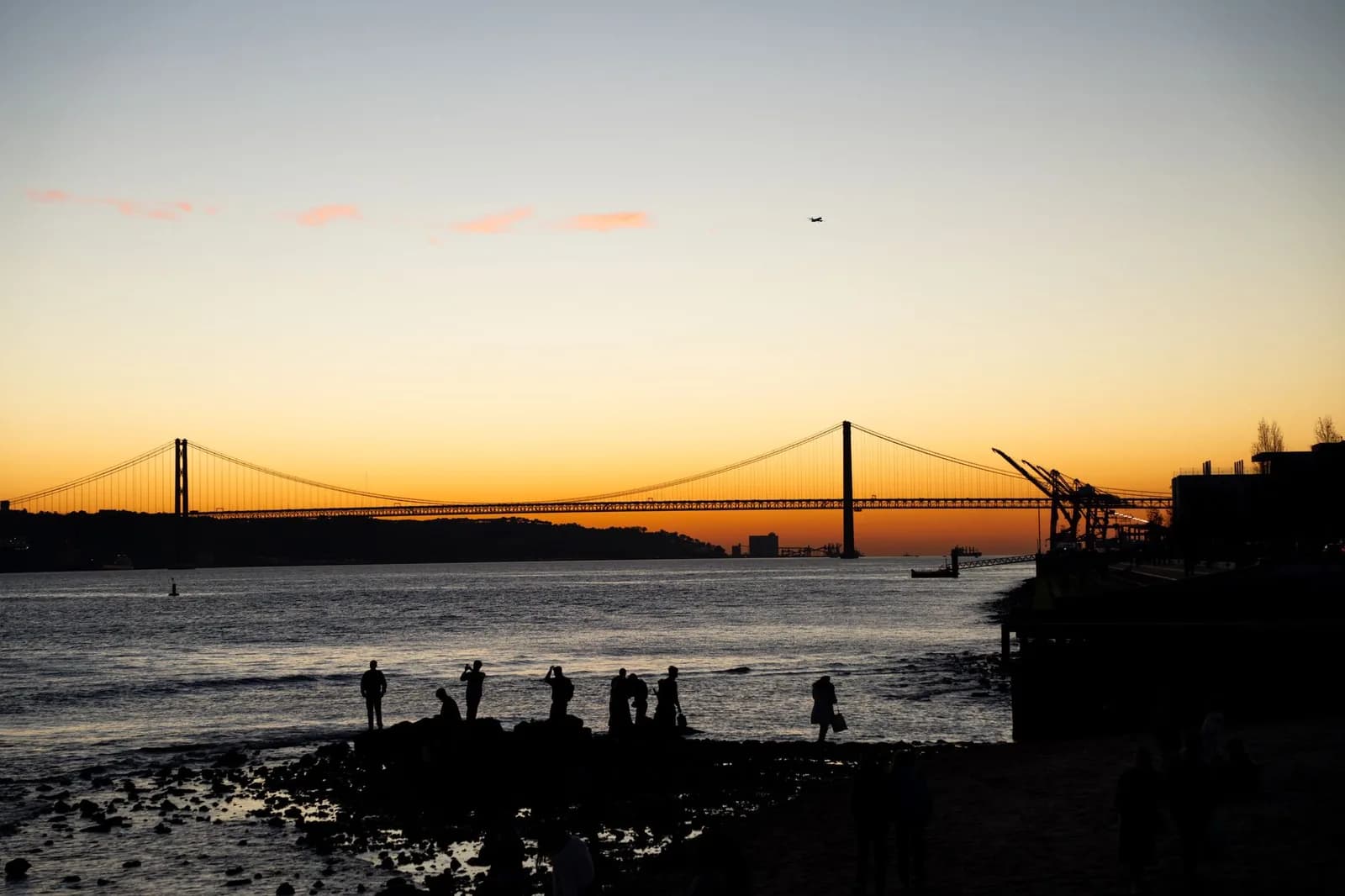 Le pont du 25-Avril en silhouette au coucher du soleil sur le Tage, avec des promeneurs au bord de l'eau