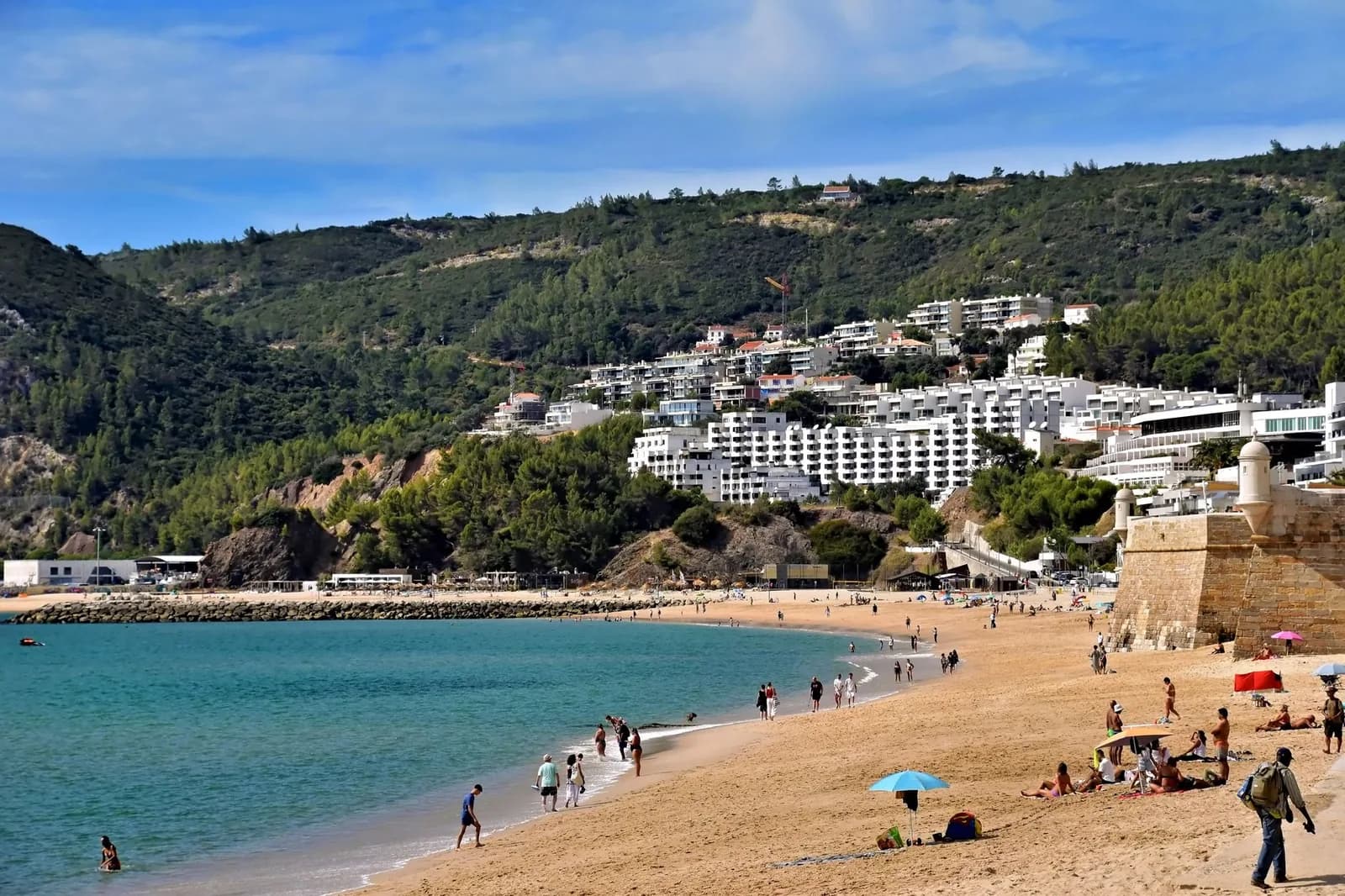 Plage de sable de Sesimbra aux eaux calmes, avec le village blanc et les collines boisées derrière