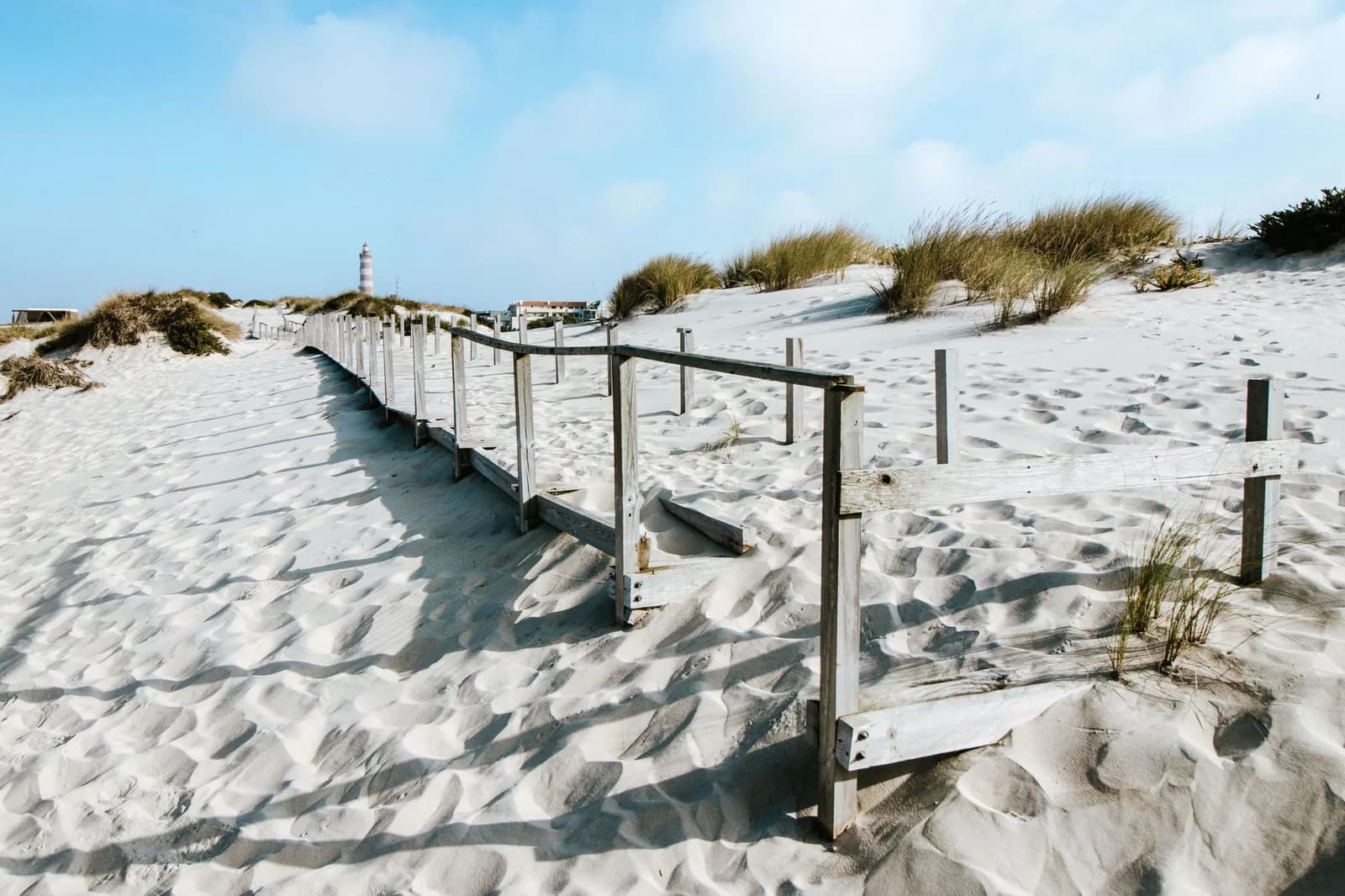 Plage sauvage de sable blanc avec dunes, passerelle en bois et phare au loin, sur la côte atlantique