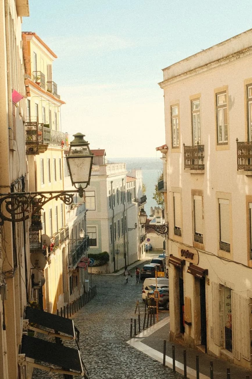 Rue en pente du Chiado bordée de façades beiges et ocre, lanterne en fer forgé et Tage au fond, Lisbonne